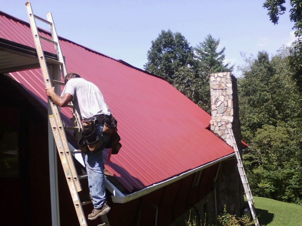 Roofer on a ladder beside a red metal roof and stone chimney — Soffit & Fascia Repair by Roofing Brothers in Gloucester